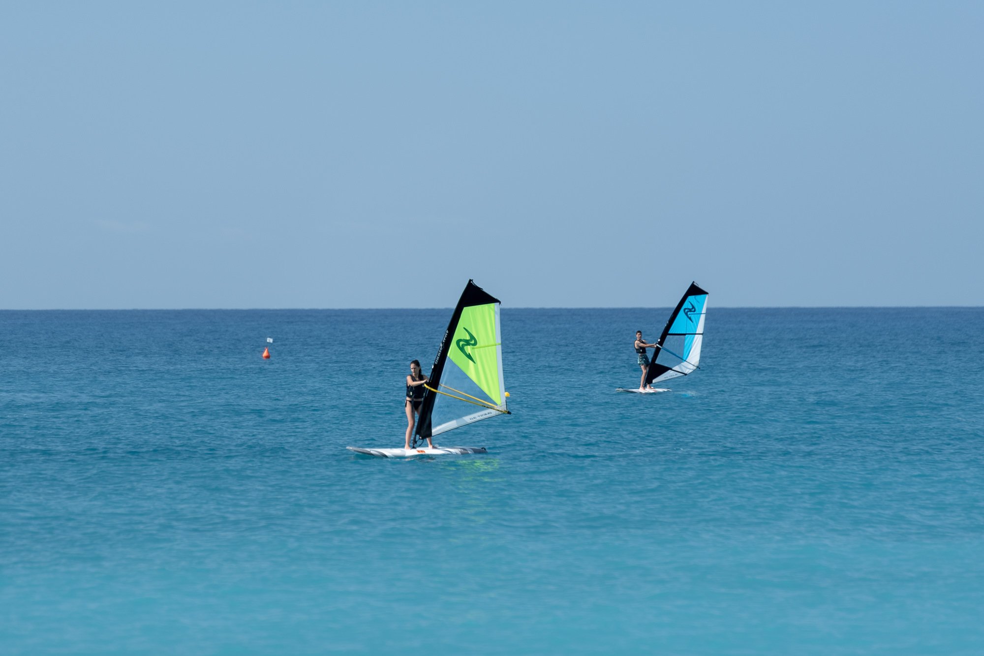 Due windsurf con vela verde e azzurra sul mare turchese di Bordighera, con una boa rossa