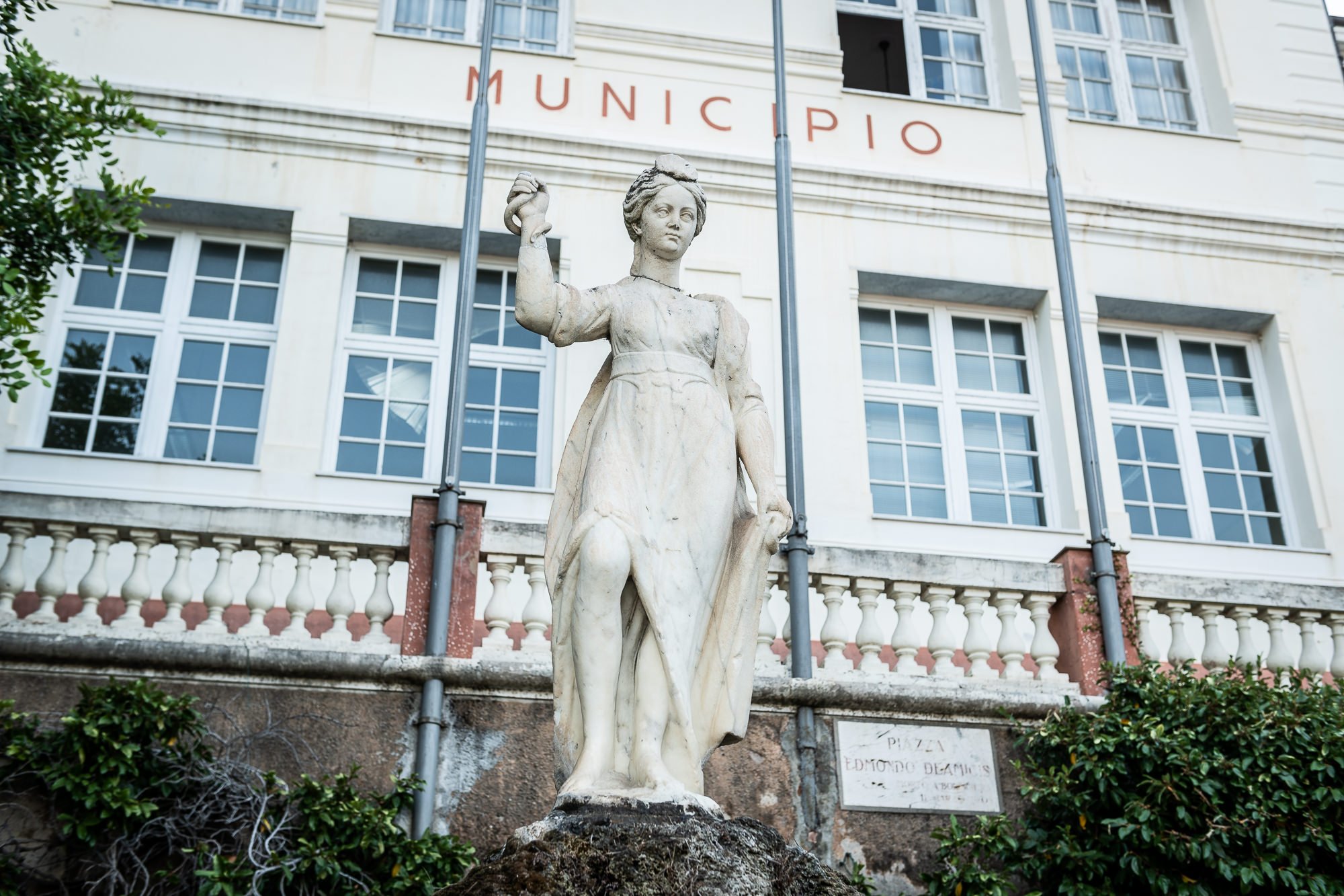 Statua in marmo bianco della Magiargè sulla fontana davanti alla facciata del Municipio di Bordighera, tra aste portabandiera