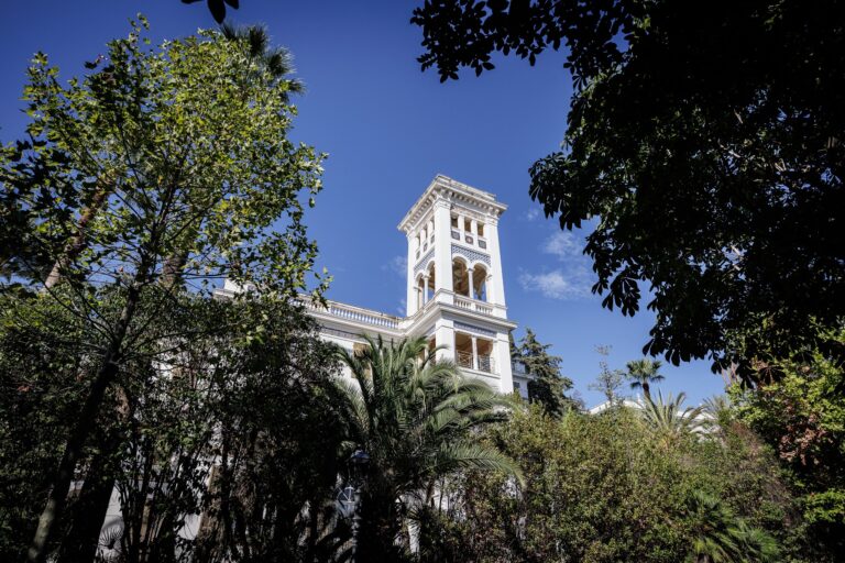 La torre bianca di Villa Etelinda che emerge tra le chiome degli alberi e le palme del parco, sotto un cielo blu intenso