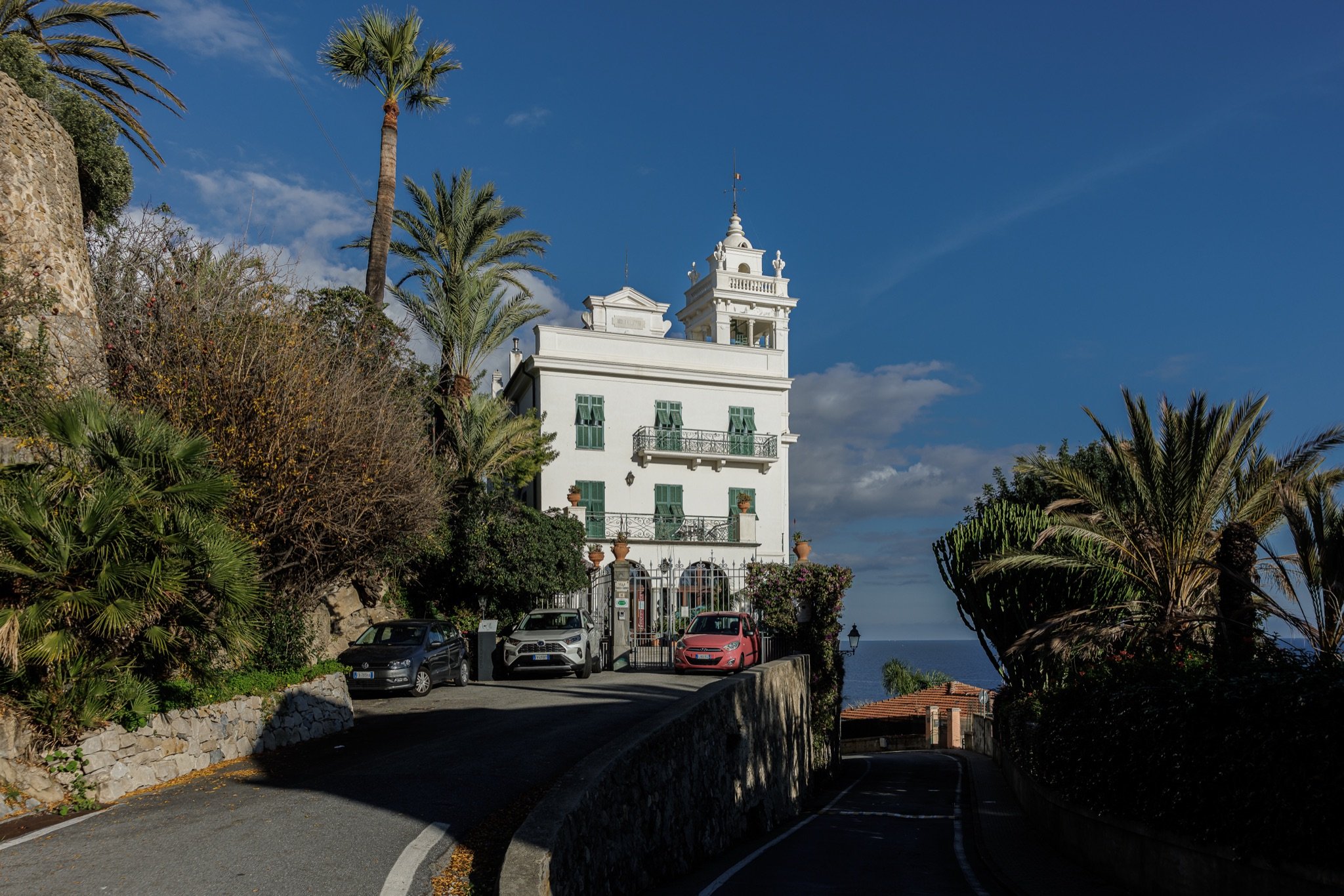 Facciata bianca di Villa Garnier con la torre e le persiane verdi, sopra la strada in salita tra palme