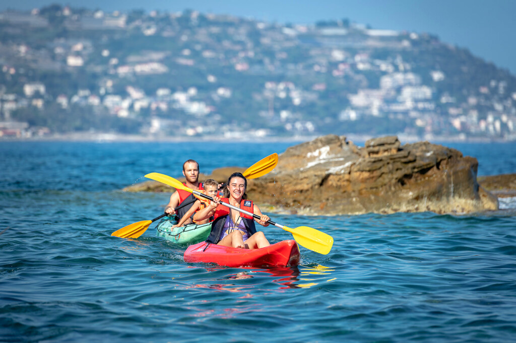 Mamma sorridente in kayak rosso e papà con bambino in kayak verde accanto a un grande scoglio di arenaria a Bordighera